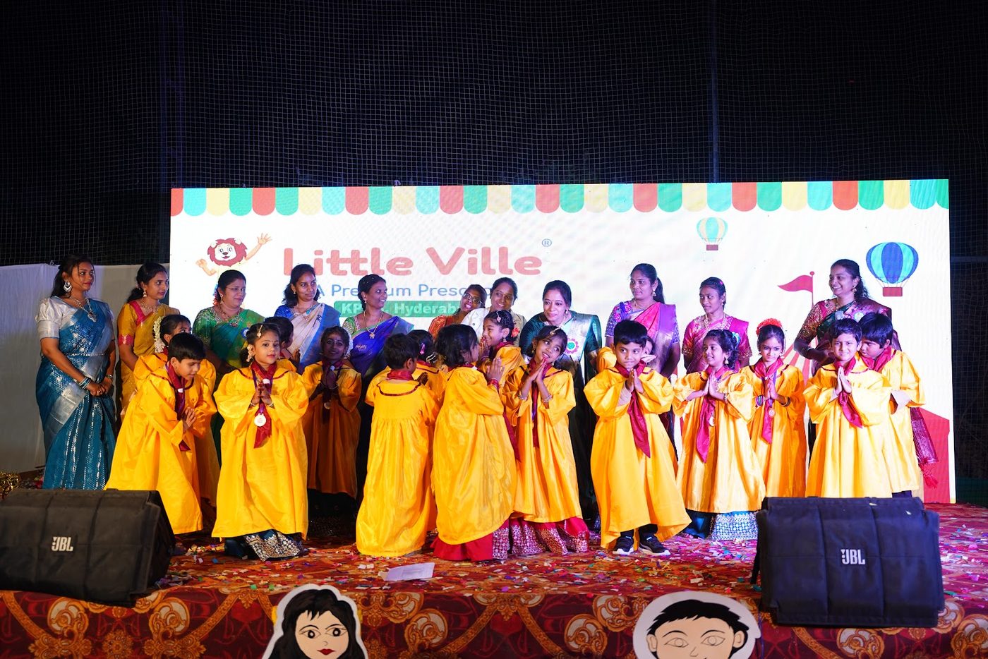 Little Ville graduates in yellow gowns celebrating on stage with their teachers at Grad Day